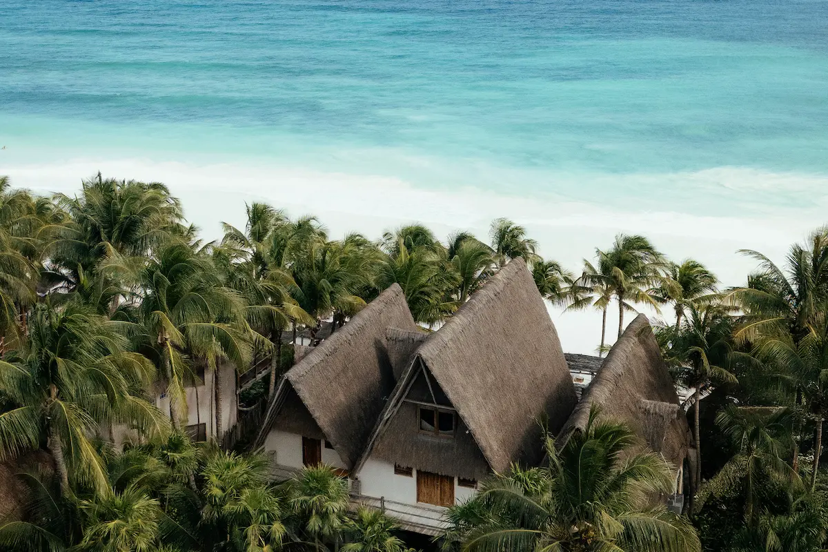 Aerial view of La Valise Tulum hotel featuring traditional thatched roofs nestled among palm trees by the turquoise Caribbean Sea in Mexico.