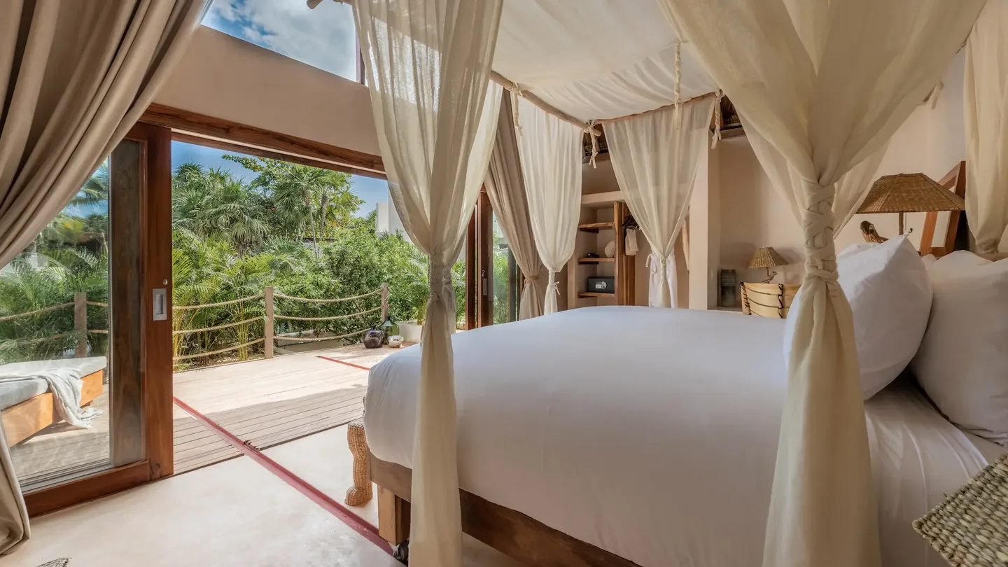 Habitación de hotel de lujo en La Valise Tulum con una cama con dosel blanca y grandes puertas de vidrio con vista escénica a la selva.