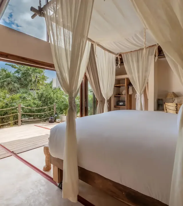 Habitación de hotel de lujo en La Valise Tulum con una cama con dosel blanca y grandes puertas de vidrio con vista escénica a la selva.
